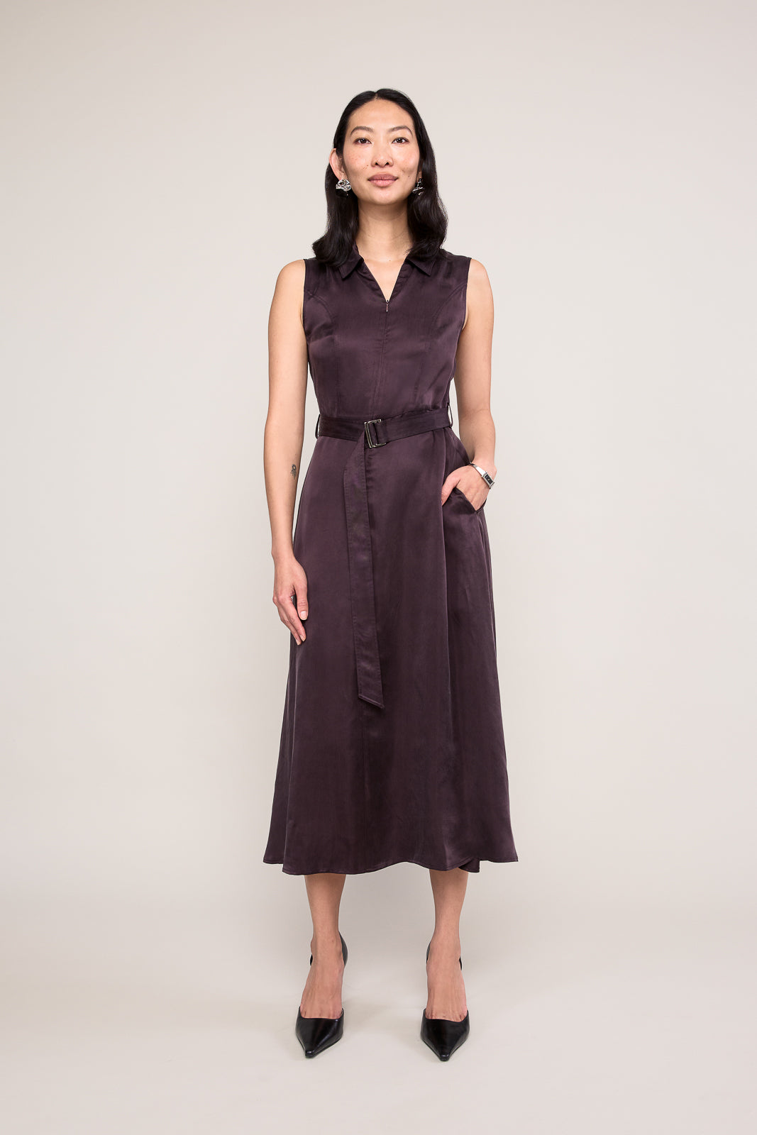 A woman modeling a sleeveless, dark plum midi dress with a collar, matching tie belt, and side pockets, paired with black pointed-toe heels, against a plain light gray studio background.