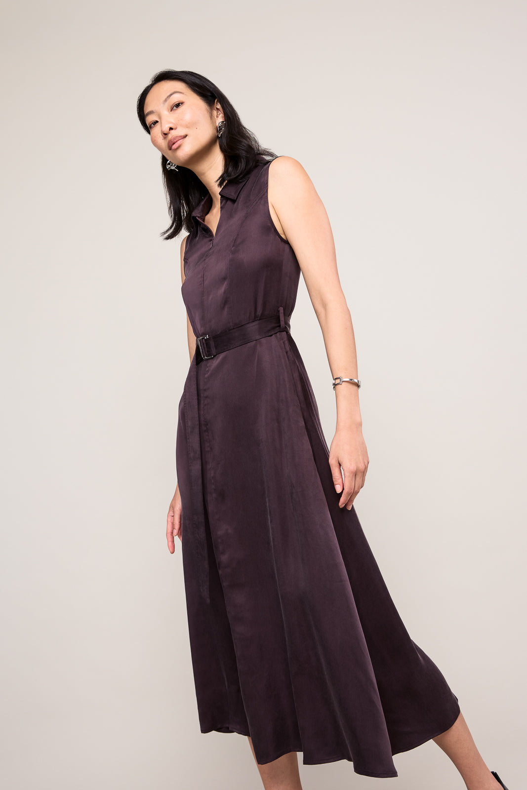 A woman modeling a sleeveless dark plum midi dress with a matching waist belt, photographed against a plain light gray studio background.