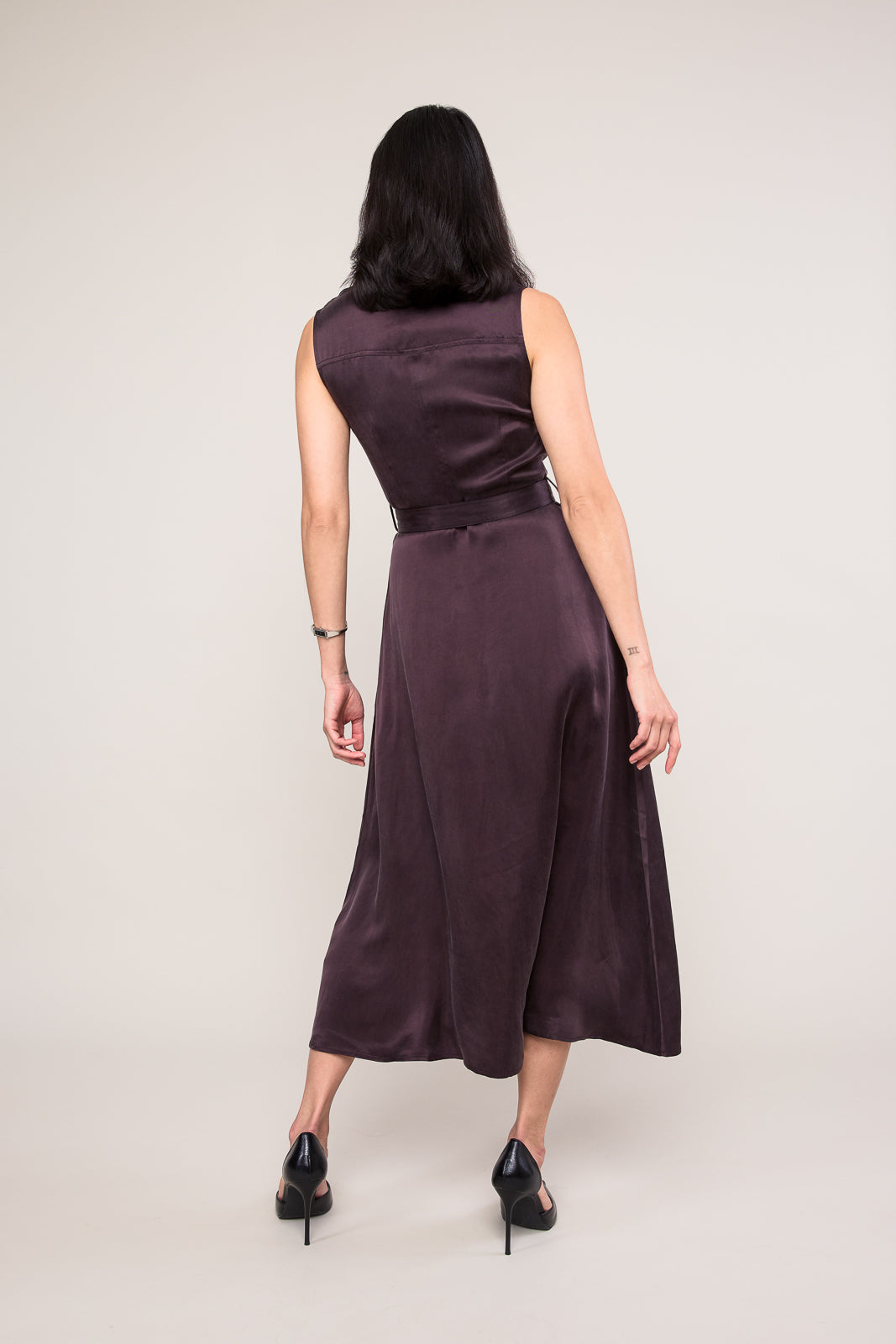 A woman viewed from the back wearing a sleeveless, midi-length dark plum satin dress with a matching belt, paired with black high heels, against a plain light gray studio background.