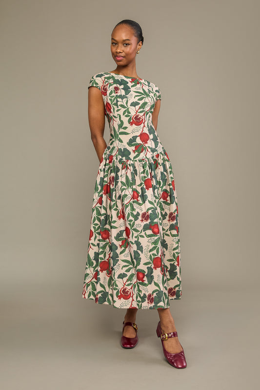 A woman modeling a knee-length cream dress with a green and red floral print, featuring cap sleeves and a gathered waist, paired with burgundy Mary Jane shoes against a solid gray studio background.