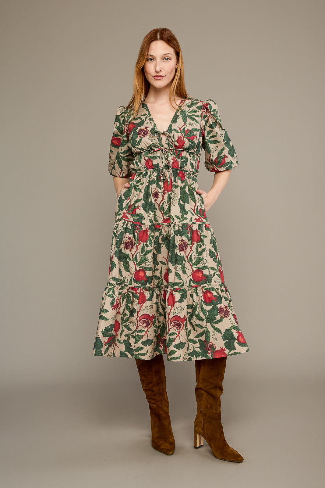 A woman modeling a knee-length floral dress with puff sleeves and a smocked waist, paired with brown knee-high boots, against a plain taupe studio background.