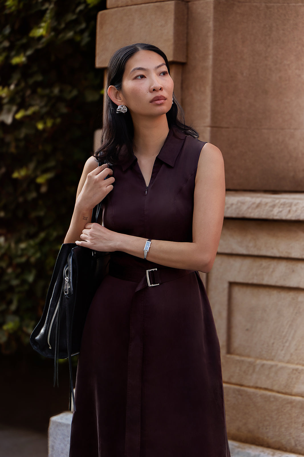 A woman wearing a sleeveless dark plum-burgundy collared dress with a matching belt and silver buckle, standing outdoors against a tan stone building wall.
