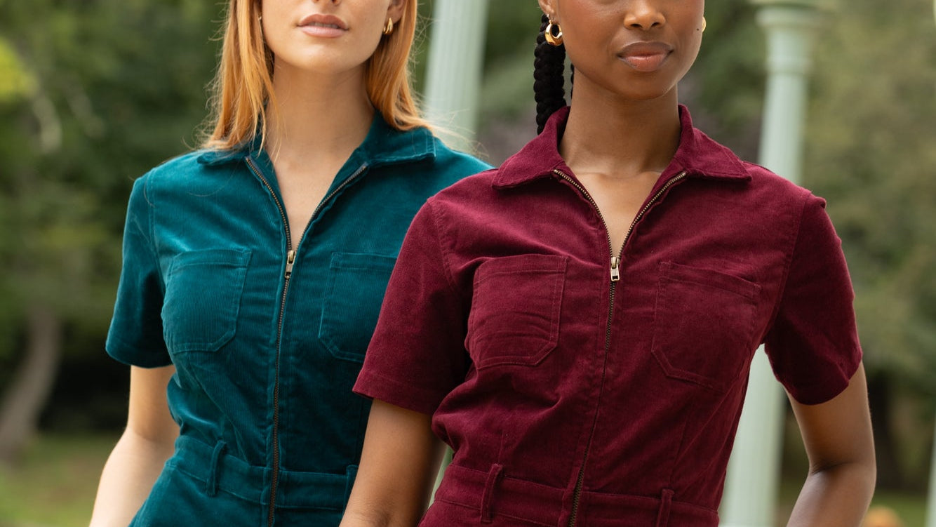 Two models wearing corduroy jumpsuits in teal and burgundy, standing outdoors in front of a blurred park background with lamp posts.