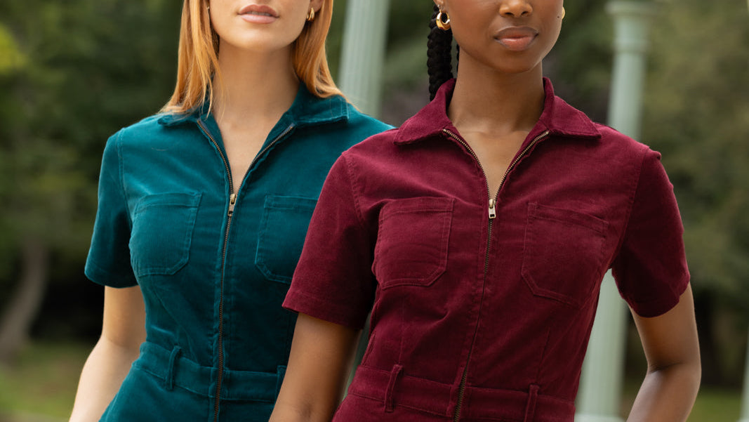 Two models wearing short-sleeve corduroy jumpsuits, one in teal and one in burgundy, standing outdoors in front of a blurred background with green trees and lamp posts.