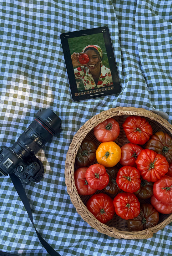 Basket of tomatoes on a checkered tablecloth with a tablet displaying a person holding a tomato.